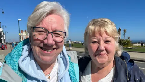 Sandra and Lynn smile at the camera as they stand near Front Street in Seaham. Sandra is dressed in a light blue hoodie and is wearing glasses with a navy frame. Lynn has a navy blue coat on.