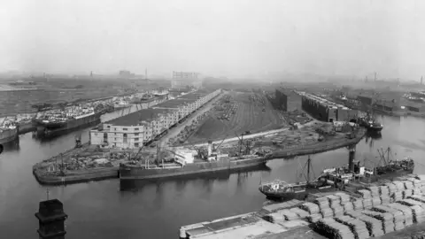 Black-and-white aerial photograph of Salford Docks showing multiple cargo ships moored along the quays, large stacks of goods in the foreground, and rows of warehouses extending into the industrial landscape under a hazy sky.