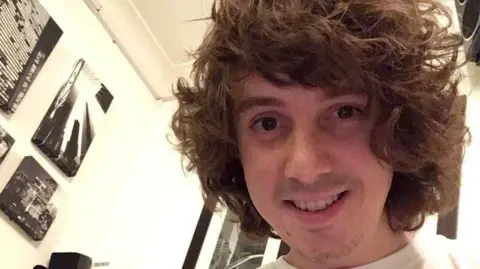 Family handout Oliver Robinson with medium length, curly brown hair poses for a selfie in what appears to be a bedroom with black and white cityscape photo canvases on the wall. He is smiling at the camera.