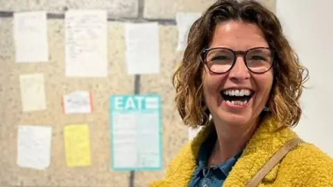 Woman laughing with chin-length wavy hair and glasses wearing a mustard yellow coat with a denim shirt and lists on a board behind.