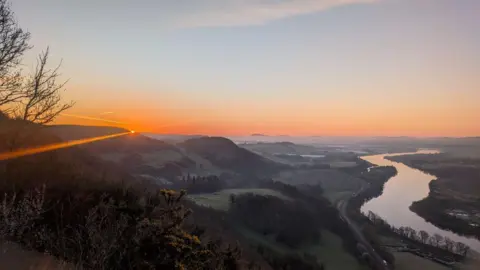 Rich Garside Organe sunrise glow appearing over hilltops in the distance, with a winding river and field and trees also in the shot. 