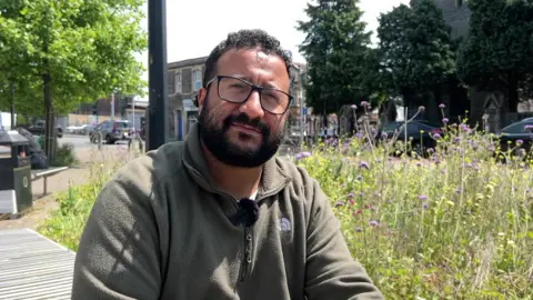 Courtney Young sitting on a bench nearby a road with a patch of grass and some flowers. He is wearing a khaki green fleece and has black rimmed glasses and curly hair.