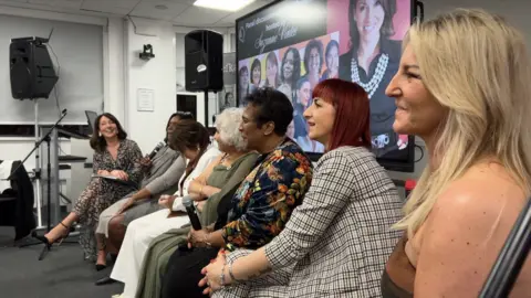 Host Suzanne Virdee sat at the far end of a panel at the Queens of the City event in Walsall, with six women sat in a row sharing their stories. The women have different coloured hair, from brown and red to blonde and black. They sit in front of a screen showing their faces.