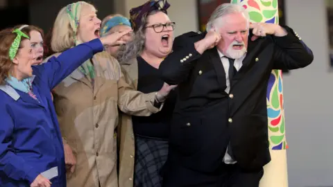 Sartorial Pictures A scene from Scottish Opera's production of Carmen, with several women singing/shouting and pointing at a man in a suit, who has his fingers in his ears as if blocking them out.