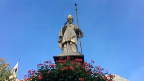 Looking up at the statue of Saint Lull as a bishop from the ground, so he is looking down at the camera. The figure is great, with bits of gold and he holds a staff. Blue sky behind and red flowers circle the bottom.