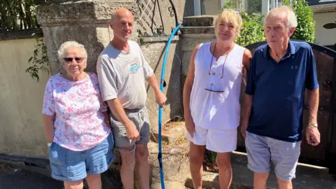 Four residents stand outside a house. There are two women and two men. They are all wearing shorts and t-shirts. One of the men is holding a blue pipe in his hand. Behind them is the stone wall of a house.