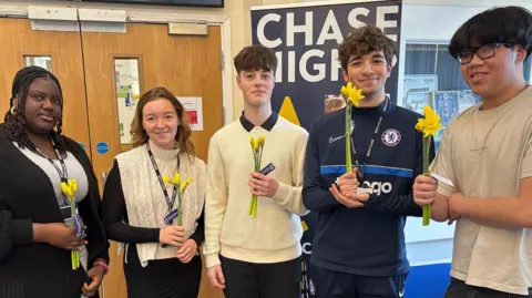 Discovery Educational Trust Five teenagers - two females and three males - stand side by side, inside in a school setting. They each hold a small bunch of daffodils. They are generally smiling.