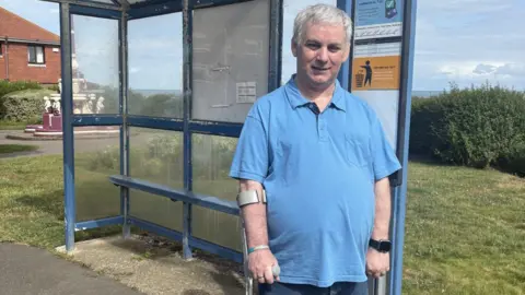 A man in his 60s wearing a blue shirt stands in front of a bus stop staring into the camera. He has a crutch in his right hand.