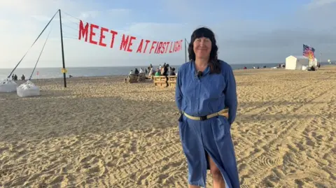 Zoie O'Brien A woman in a blue dress smiles at the camera, her hands behind her back. She has black long hair and is standing on a sandy beach, the sea behind her and a banner stretched between poles which reads MEET ME AT FIRST LIGHT 