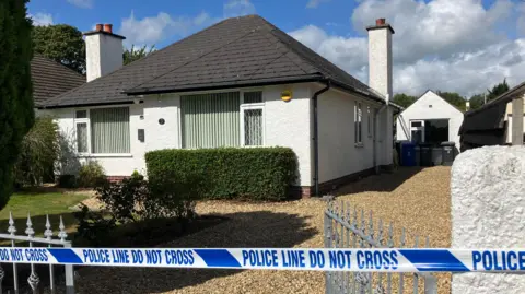 A white bungalow with a black roof with a garden in front of it and a stoned driveway is cordoned off with blue police tape.