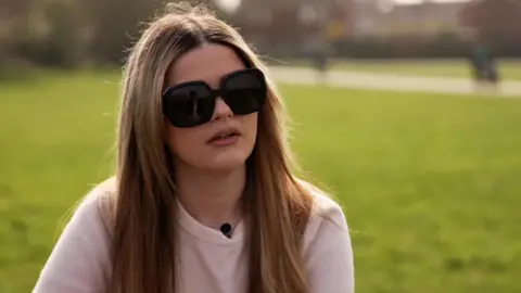 Annabelle Mackay is sitting outside in a park in the sunshine. She has long hair and is wearing a pink T-shirt and large, dark sunglasses.