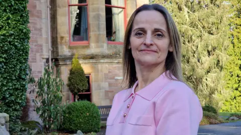 Susan has shoulder length blonde hair and wearing a pink top. She is standing outside Fairburn care home. It is a period building with trees in the ground.