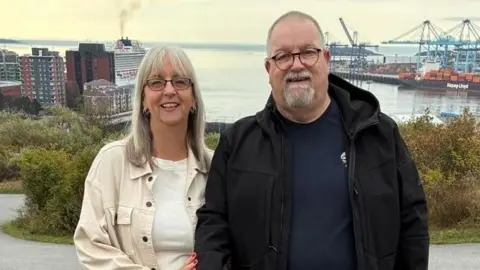 Nigel Benwell A couple in their sixties stand in front of the New York docks on an Autumn day. They are smiling to camera. She is wearing a beige denim jacket and beige t-shirt, he is in a dark rain jacket and blue t-shirt. 