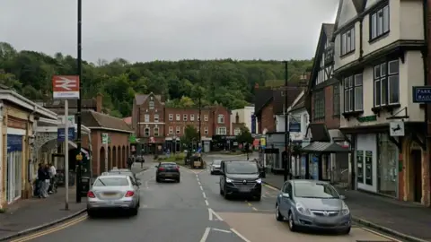 Station Avenue in Caterham has the railway station on one side and shops and residential accommodation nearby. There are cars on the road and people waiting outside the station, with countryside in the background.
