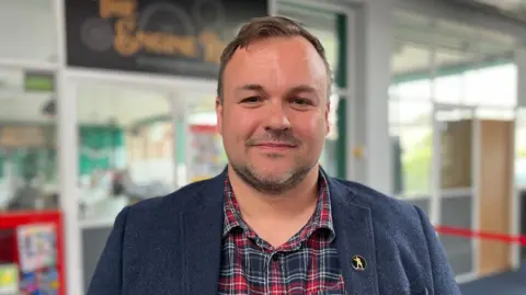 Shaun Whitmore/BBC A head and shoulders shot of Terry Jermy, who has short light brown hair and a short beard. He is wearing a mid-blue tweedy jacket over a red, blue and white tartan shirt. He is standing in a building.
