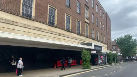 Owen Sennitt/BBC A large boarded-up department store in Norwich city centre can be seen, with people walking past on a grey cloudy day.