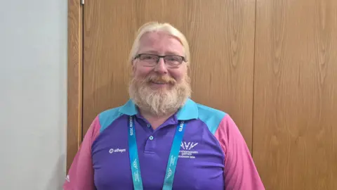 Peter Craig - a man with a fair beard, glasses and fair hair, wearing a commonwealth games uniform of purple, blue and pink.