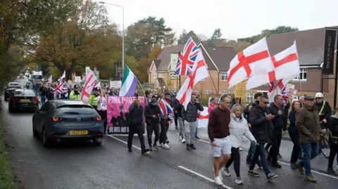 BBC / Claudia Sermbezis Hundreds of people holding St George flags and Union Jack flags marching along a road. There are cars driving by. 