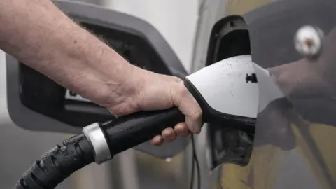 A hand holds a cable as it is plugged into a silver electric car.