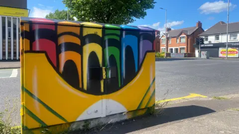 A junction box which has been painted to look like a yellow box of crayons. There are 5 crayon colours - red, orange, yellow, green, blue and purple. There is a road and houses behind.