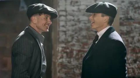 Netflix Two men (actor Barry Keoghan and Cillian Murphy) stand facing each other indoors, both wearing flat caps and period-style clothing. The setting appears industrial, with an exposed brick wall and soft, directional lighting. Their close positioning suggests an engaged conversation within a historical or dramatic scene.