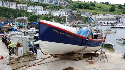 EX RNLB Guide of Dunkirk in Cornwall. The vessel is on a slipway out of the water. The vessel is white, blue and red. Pedestrians are watching as the boat is removed from the water. Boats are in the water in the background. Properties overlook the water.