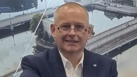 Gary Quittenton-Shaw Image of Gary. He is wearing a navy suit jacket and a white shirt. He is wearing glasses and is stood in front of 