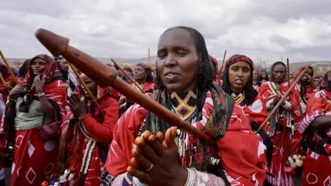 Women wearing red traditional outfits, holding a wooden stick