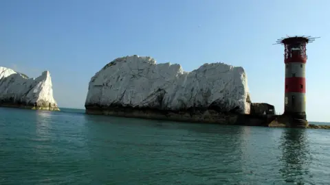 View across calm coastal waters toward large white chalk sea stacks and a striped lighthouse standing on a stone pier. The cylindrical lighthouse, painted with red and white horizontal bands, rises on the right side of the image beside a rugged chalk rock formation. Additional chalk cliffs are visible to the left, separated by a narrow channel of sea. The turquoise water reflects the structures, while a clear blue sky fills the background. Bright sunlight highlights the contrast between the white cliffs, the colourful lighthouse, and the surrounding water.