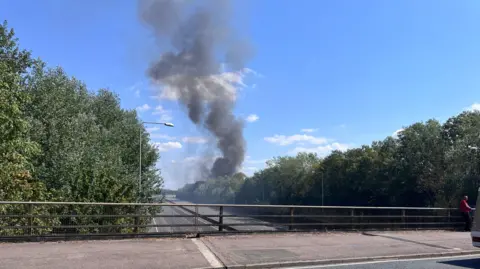 Nicola Haseler/BBC The shut M25, showing a bridge across it. Smoke is billowing up in the air and the bridge has railings on it. There are trees either side.