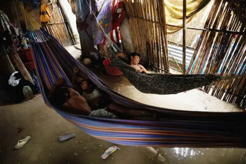 Getty images are lying in one swing, and the other in a separate swing, inside a room made of wooden poles. Under the swing, there is shallow water on the ground, in which two sandals float in water.