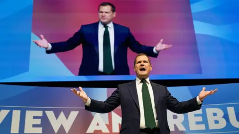 PA Media Robert Jenrick wearing a blue suit, delivering a speech on stage at the Conservative Party conference in Birmingham