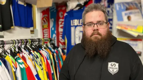 Daniel Rees lasting  beside a rack of multi-coloured shirts and successful  beforehand   of a partition  displaying squad  shirts.