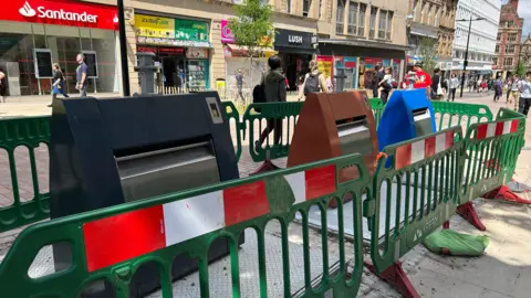 Three large bins in different colours are surrounded by plastic fencing on a high street