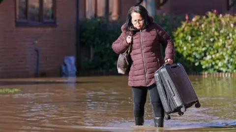 A woman in a purple padded jacket struggles with a black suitcase in flood-water. The water is up to her ankles as she wades through in wellies with a bag over one shoulder. Behind her is the wall of a house and bushes, with water covering the ground around her