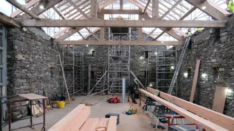 Freshfield The inside of the building during construction. The roof has been removed and new beams have been installed overhead. A workman is cutting a large wooden beam.