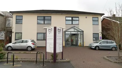 Lodge Causeway Dental Centre seen from the outside. It has a paved area in front with two cars parked there, and the building itself has cream walls and black-framed windows