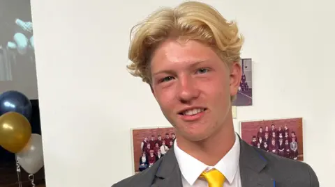 John Devine/BBC Ellis has short blonde hair and is wearing a white shirt, grey blazer and a yellow tie. He is standing in front of a display which has old school year pictures on it.