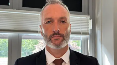 Det Con Lee Harshey-Jones. He looks at the camera with a neutral expression, and wears a white shirt, black suit jacket and bronze tie. He has short grey hair and short stubble.