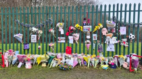 Paul Dempsey / Rothwell Juniors FC Bouquets of flowers, soft toys, pictures and cards placed up against a green metal fence outside a football club.