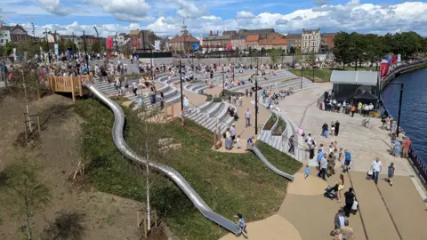 LDRS A crowd gathers in a modern outdoor amphitheatre beside a waterfront. People are seated on tiered stone seating and standing in the central plaza area during a community event beneath dramatic cloud formations.