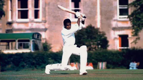 Lansdown Cricket Club A man in cricket whites swings a cricket bat over his head while lunging forwards on his left knee.