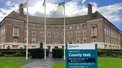 Somerset Council County Hall in Somerset. It is a brown brick, four-storey building with lots of windows and three flags outside. A turquoise sign reading "welcome to County Hall" is outside the building.