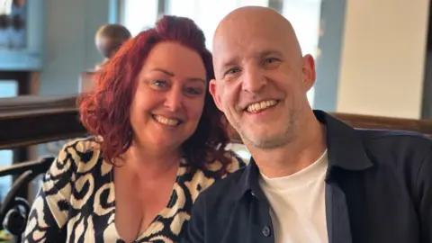 Phil Mansford Nicole and Phil Mansford, sitting down, smiling at the camera. Nicole has long red hair, wearing a black and white dress, Phil has a bald head, grey goatee, an earing in one ear and is wearing a white T-shirt and black shirt.