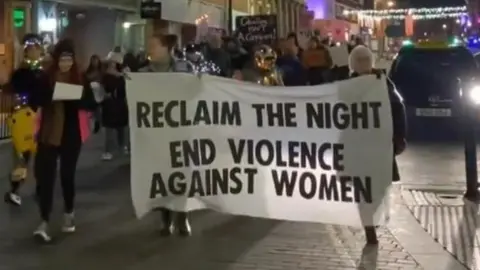 A group of women marching through Northampton in 2023. The two front women are holding a big white banner which says reclaim the night end violence against women written in black letters