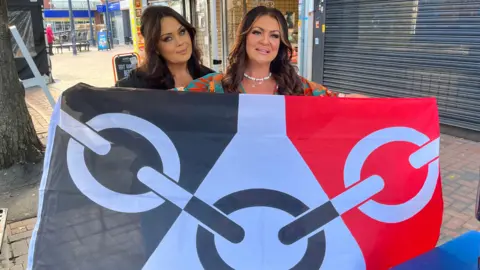 BBC Two women stand on a street holding the flag of the Black Country. The flag is black, white and red with chain representing the region's manufacturing heritage.