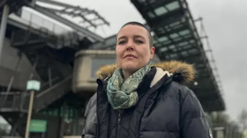 Campaigner Jo Howell stands in front of the National Glass Centre on the banks of the River Wear, wearing a thick waterproof jacket and a scarf. She has short, dark hair. 