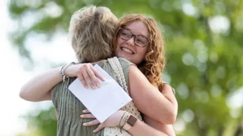 A girl with curly red hair and glasses hugs a woman with short blonde hair. The girl is smiling and holding a white piece of papers. You can't see the other woman's face but she wears a green top and beige bag. Behind them are green trees.