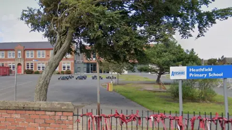 Google Healthfield Primary School is a red brick, two storey building with a large playground in front of it. The playground has several picnic tables with some patches of grass with a couple of trees. A blue sign reads "Heathfield Primary School". 