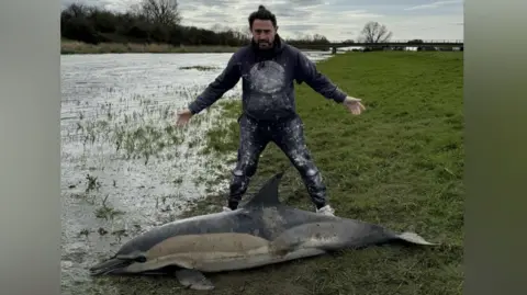 Archie Wayman Glen Wayman is standing behind a dolphin which is on the floor next to a riverbank. Wayman is wearing black trousers and a jumper.